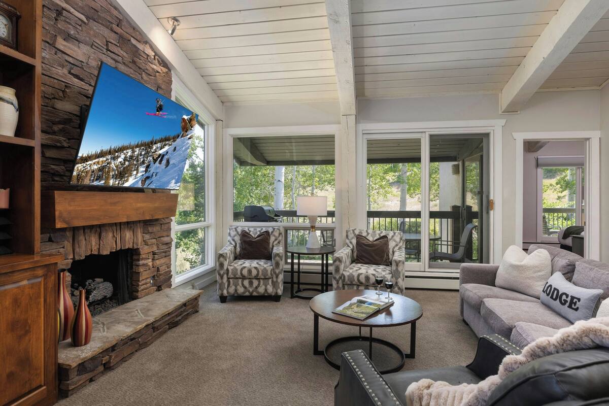 A cozy living room with a stone fireplace, comfortable seating, and a large TV screen displaying a snowy scene.