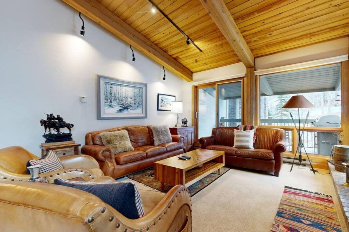 A spacious living room with a wooden ceiling, leather sofas, a coffee table, and a large window providing a view of the outdoors.