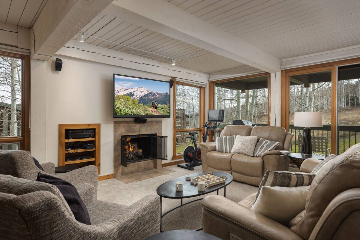 A spacious living room with a fireplace, comfortable seating, a television, and large windows offering a view of the outdoors.