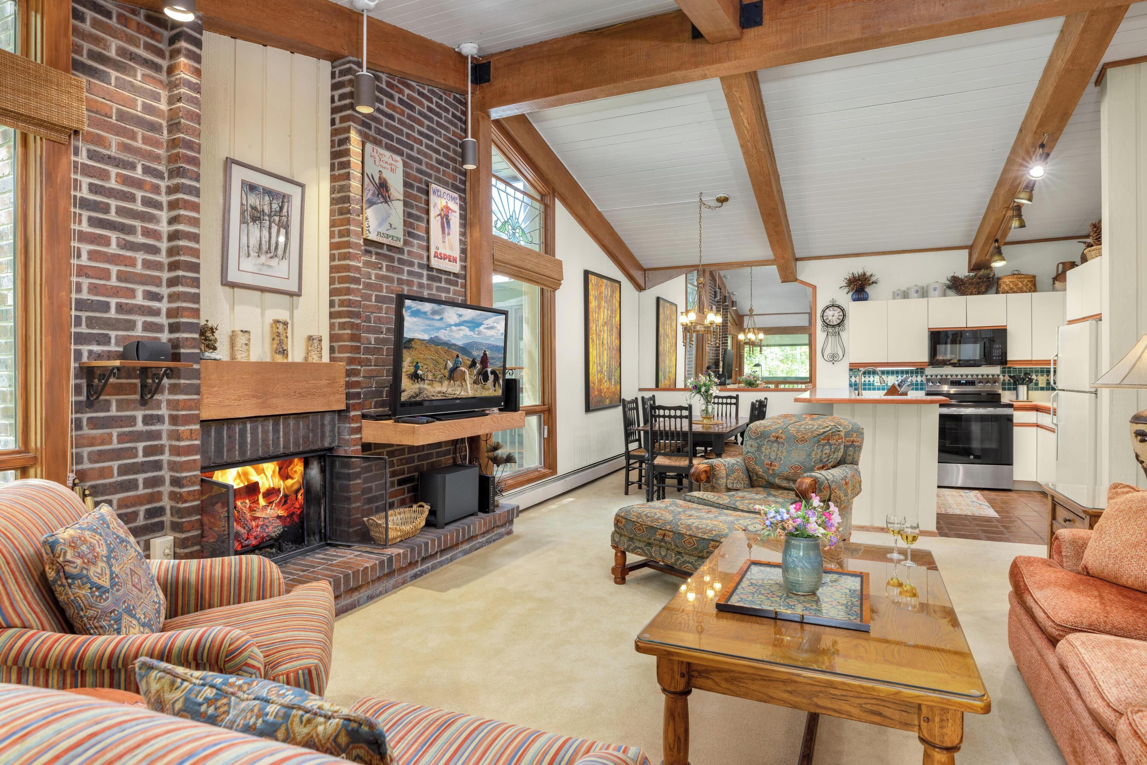 A spacious living room with a fireplace, comfortable seating, and a kitchen area in the background.