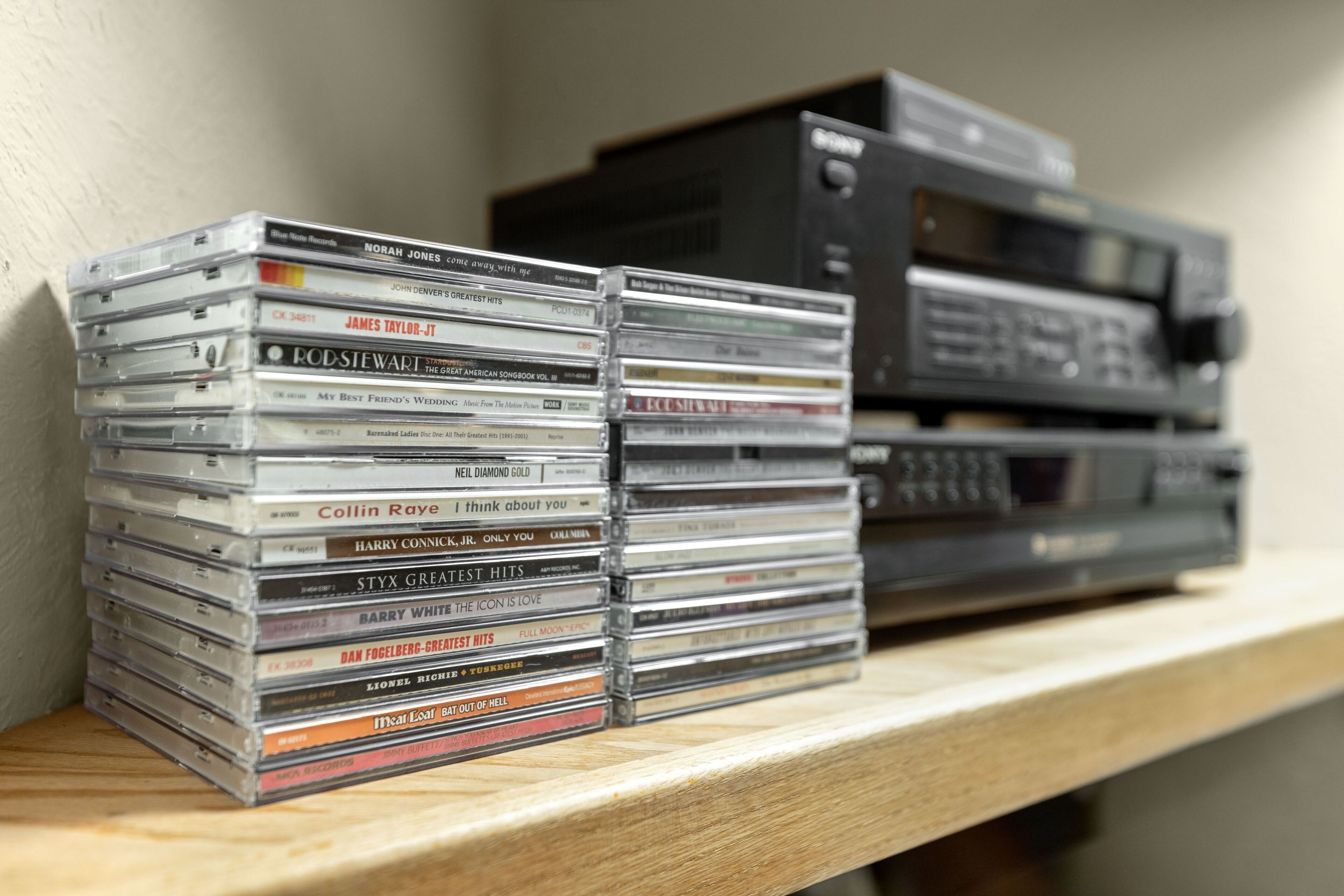 A stack of compact discs on a wooden shelf next to a black electronic device.