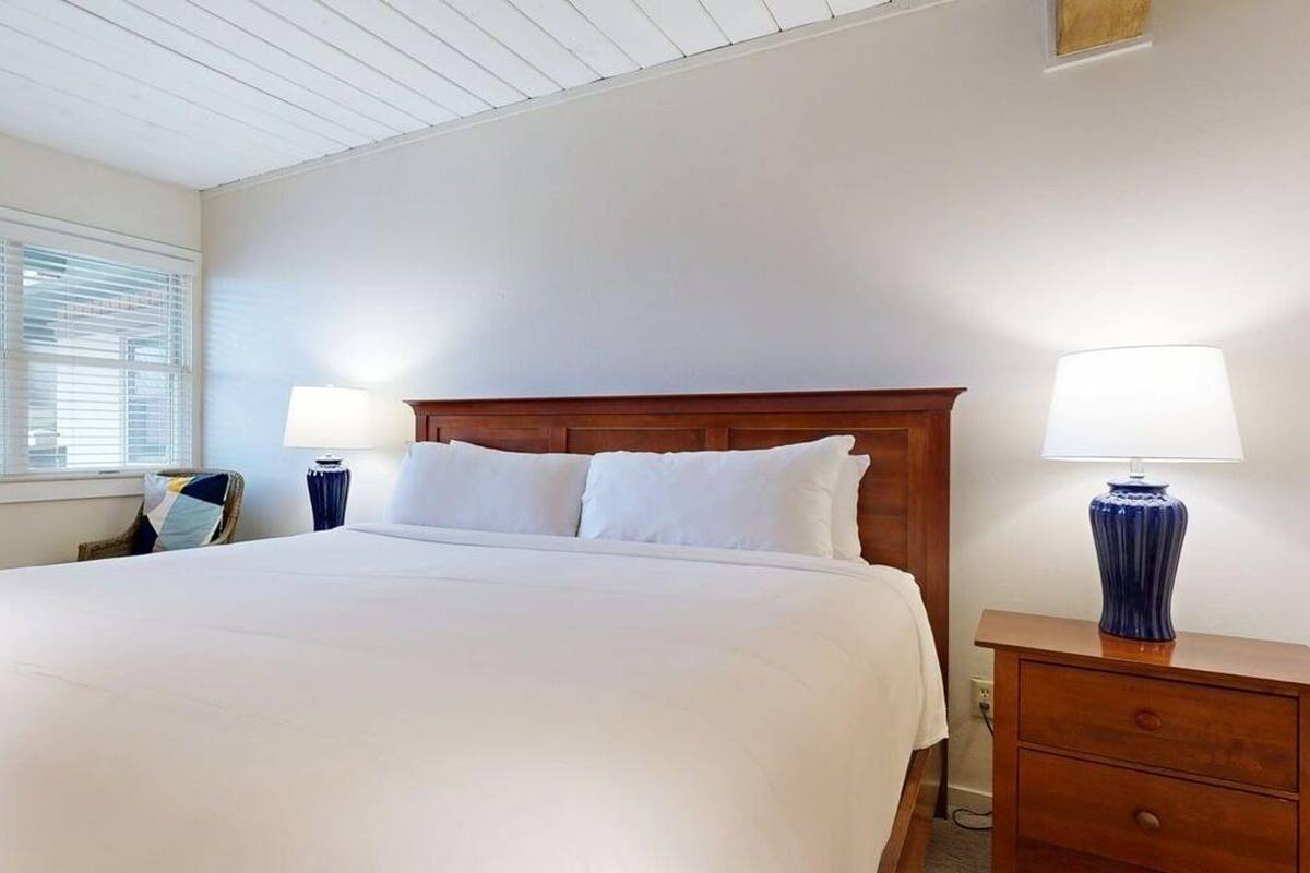 A neatly made bed with white linens in a room with white walls and a wooden headboard. There are two bedside lamps and a small wooden nightstand with a lamp.