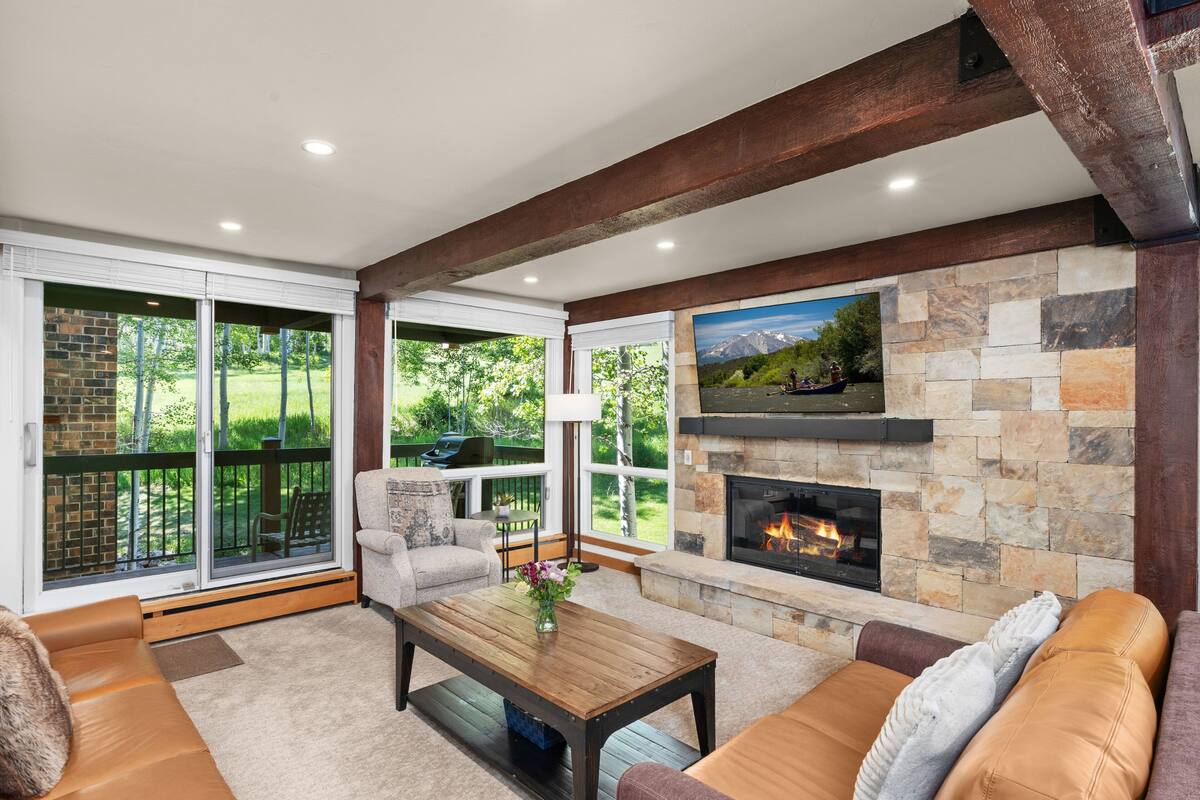 A cozy living room with a fireplace, large windows, and a stone accent wall.
