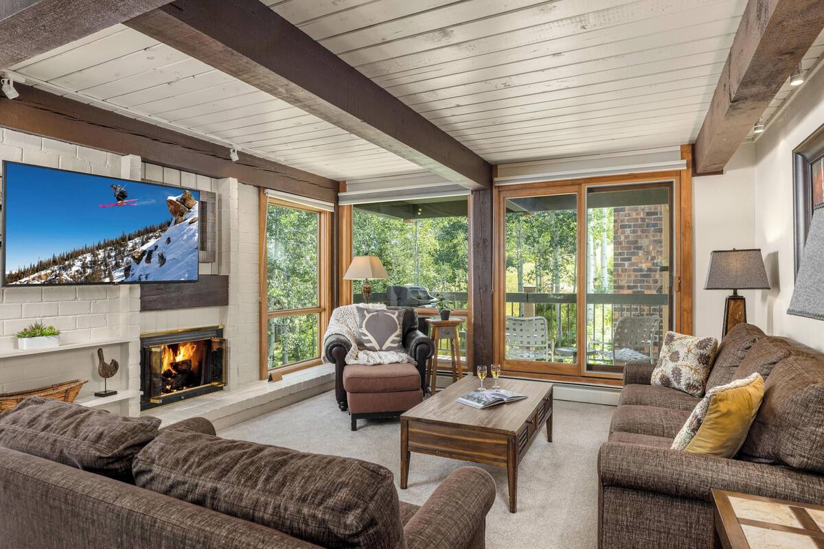 A cozy living room with a fireplace, comfortable seating, and large windows offering a view of the outdoors.