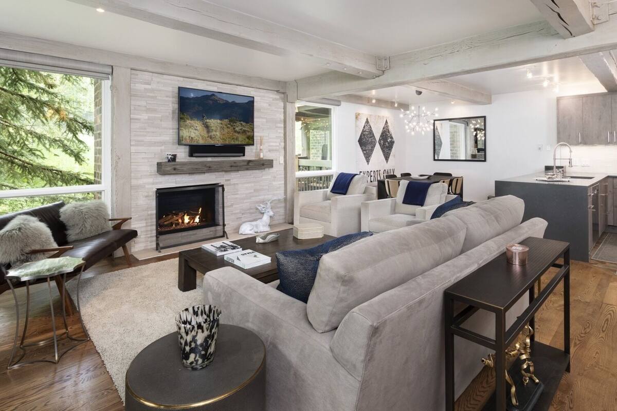 A modern living room with a fireplace, a large sofa, a coffee table, and a kitchen area in the background.