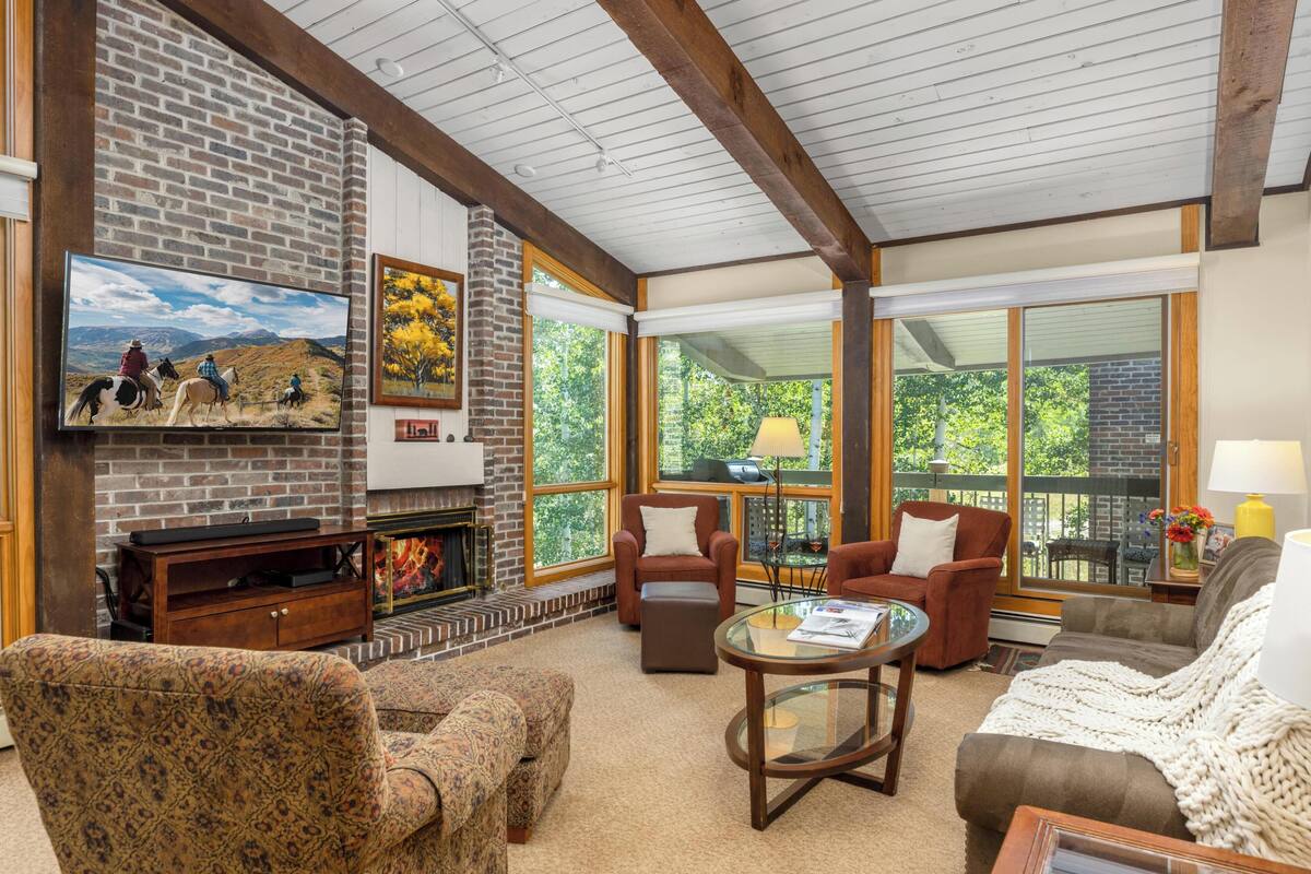 A cozy living room with a fireplace, comfortable seating, and large windows offering a view of the outdoors.