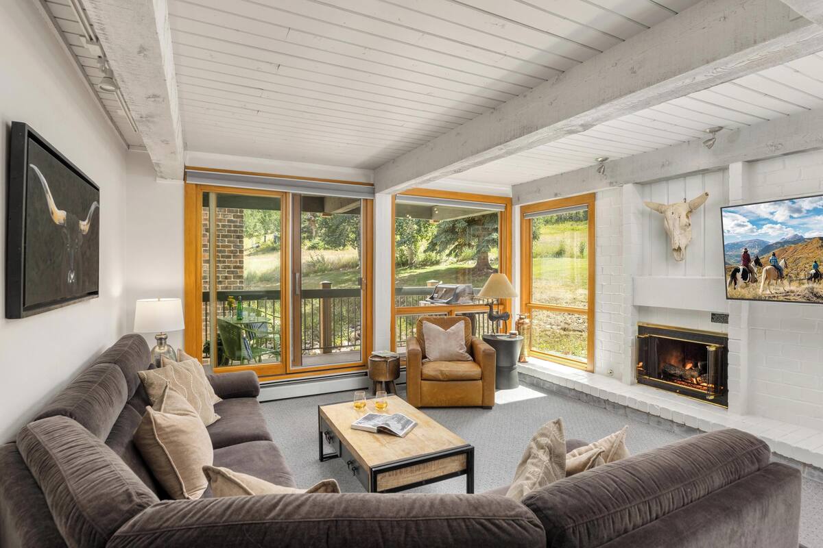 A spacious living room with a grey sofa, a wooden coffee table, and a fireplace. Large windows offer a view of the outdoors.