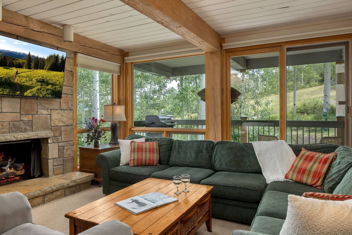 A cozy living room with a stone fireplace, green sofa, wooden coffee table, and large windows offering a view of a green landscape.