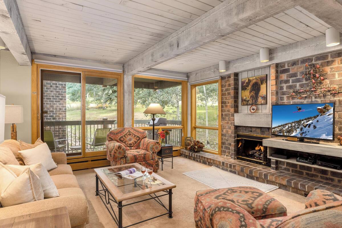 A cozy living room with a fireplace, comfortable seating, and large windows offering a view of the outdoors.