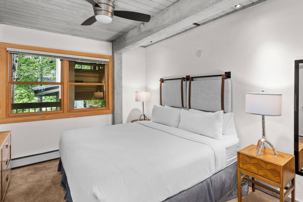 A bedroom with a large bed, a wooden nightstand, a lamp, and a window with a view of greenery.