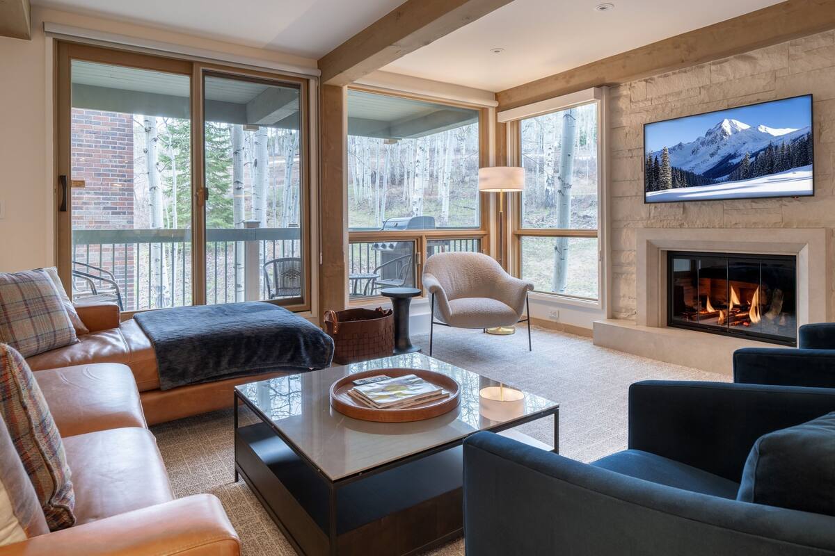 A spacious living room with a fireplace, large windows, and a television mounted on the wall.