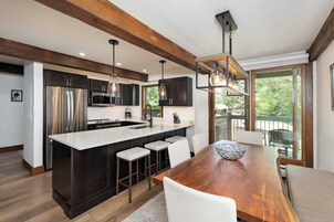 Una cocina moderna con una isla central, muebles oscuros, electrodomésticos de acero inoxidable y un comedor con mesa de madera y sillas blancas.