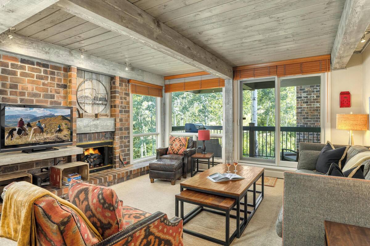 A cozy living room with a fireplace, comfortable seating, and large windows offering a view of the outdoors.