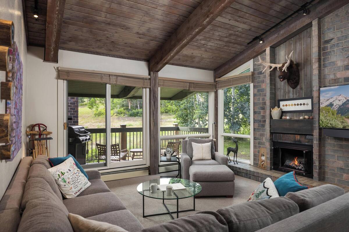 A cozy living room with a fireplace, a sofa, a glass coffee table, and a view of the outdoors.