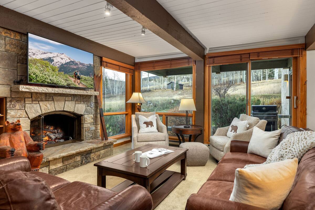 A cozy living room with a stone fireplace, leather furniture, and large windows offering a view of a mountainous landscape.