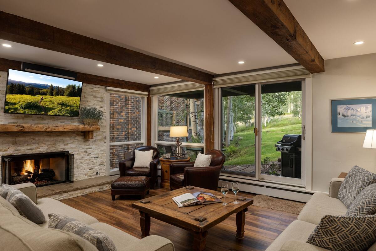 A cozy living room with a fireplace, comfortable seating, a television, and large windows offering a view of the outdoors.