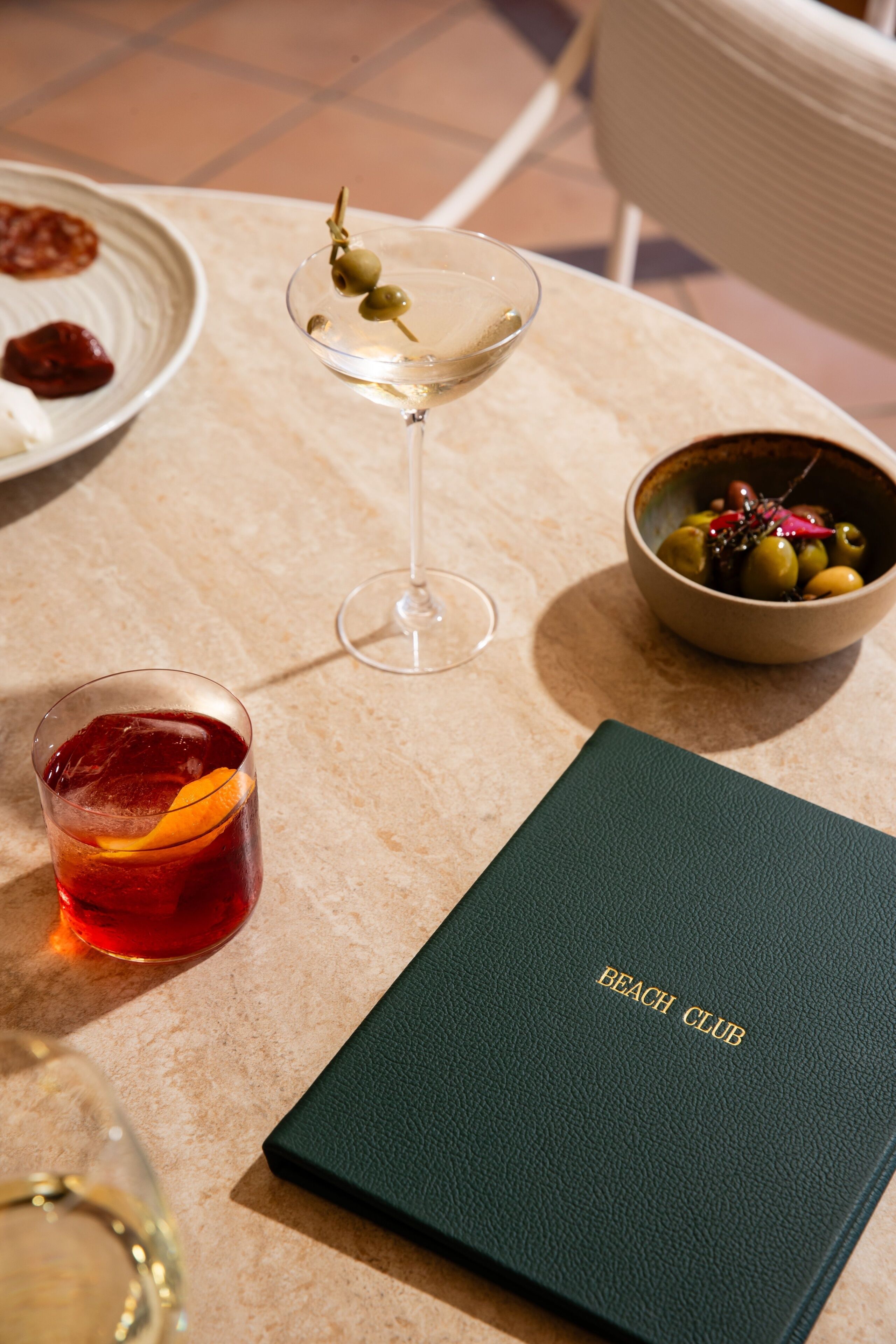 Een tafel met een martini glas, een cocktail met een oranje schijfje, een schaal olijven en een menukaart met de tekst 'BEACH CLUB'.