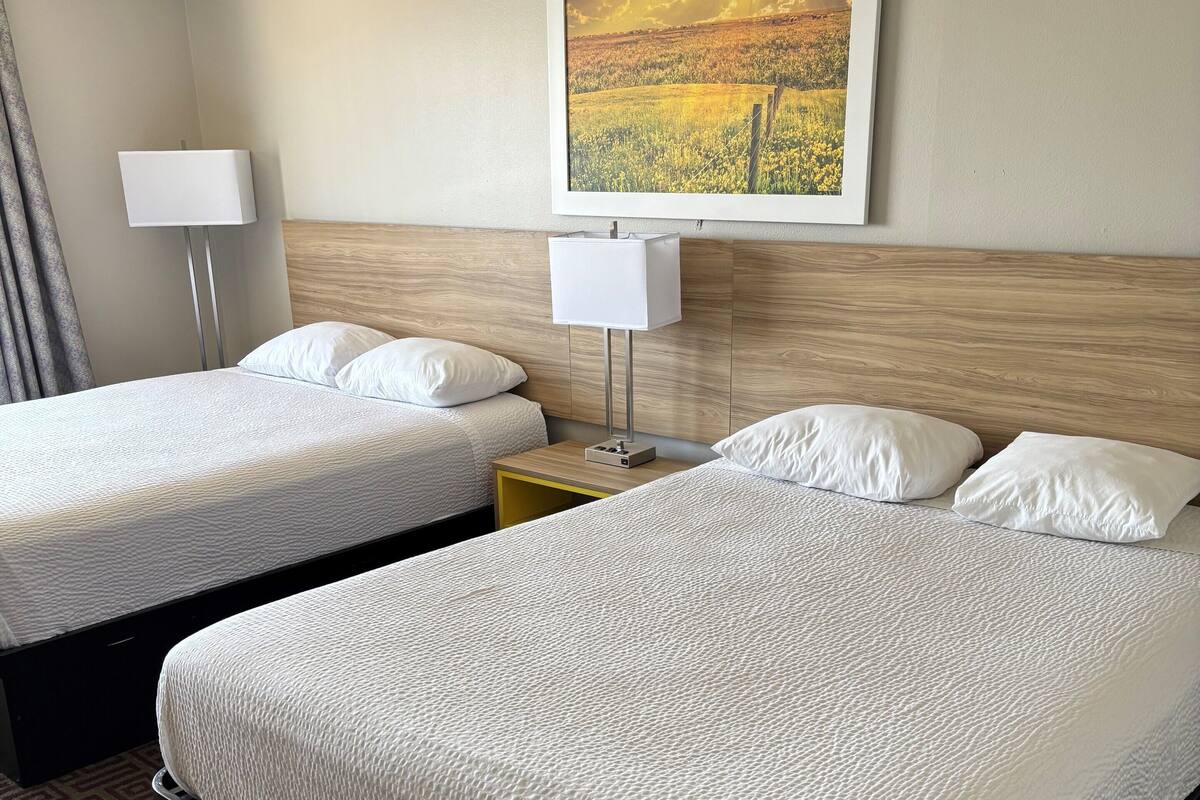 A hotel room with two beds, a nightstand with a lamp, a painting on the wall, and a patterned carpet.