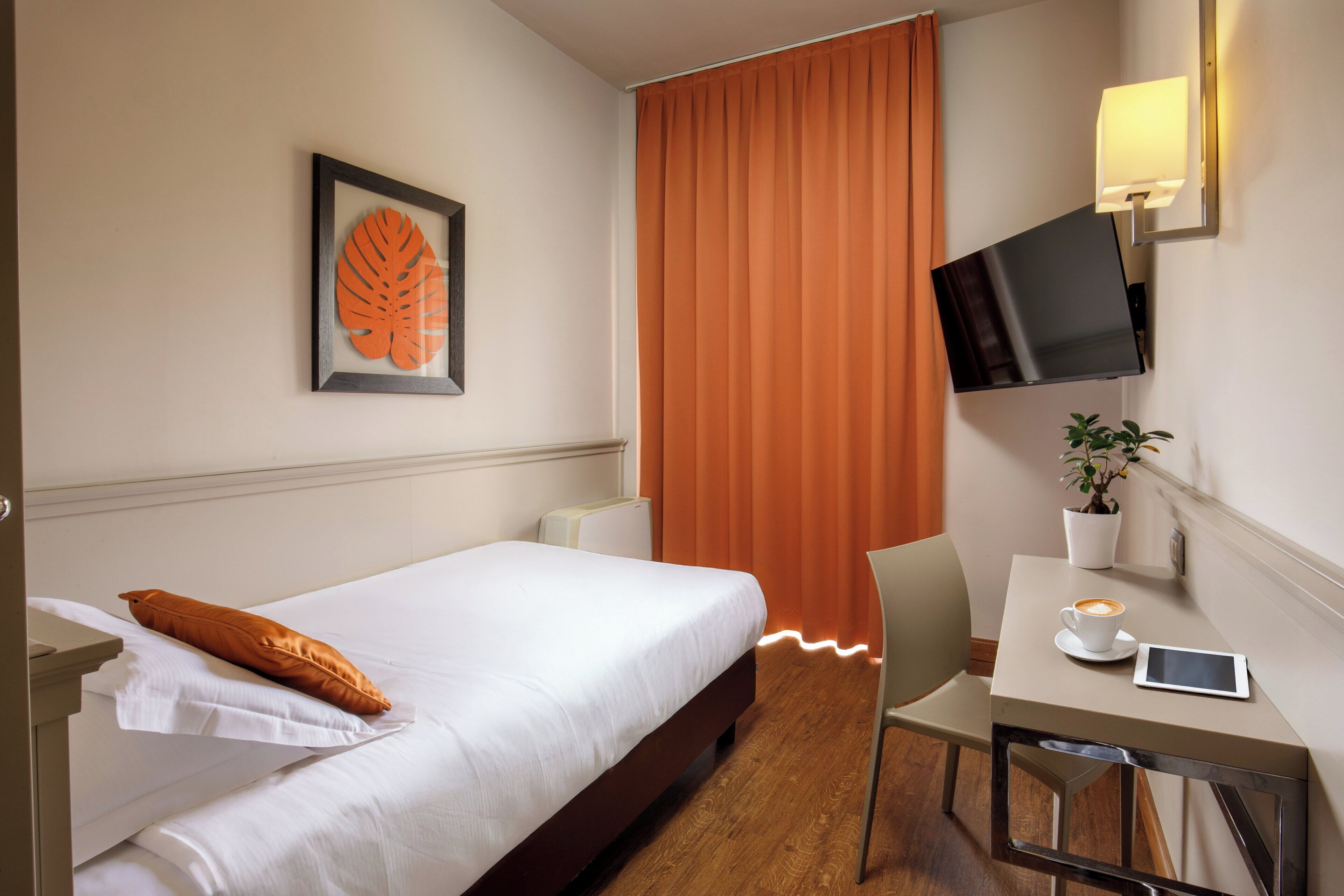 Habitación de hotel con una cama, un escritorio con una taza de café, una tableta, un televisor y una planta.