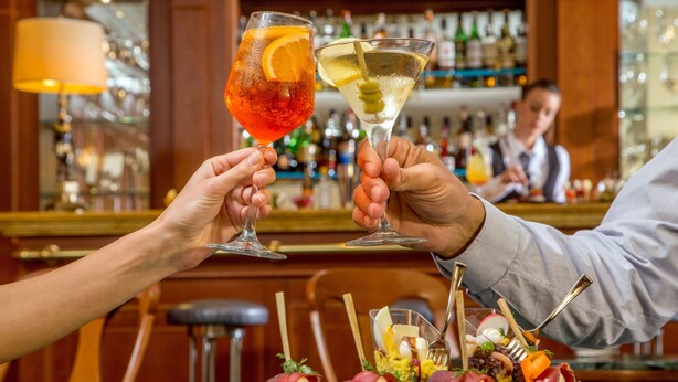 Un bar con dos personas brindando con bebidas, una barra y una variedad de botellas al fondo.