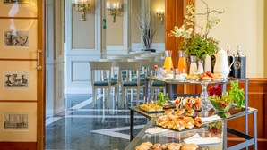 Una mesa de buffet con distintos alimentos y bebidas, un expositor de vidrio y un jarrón decorativo con flores.