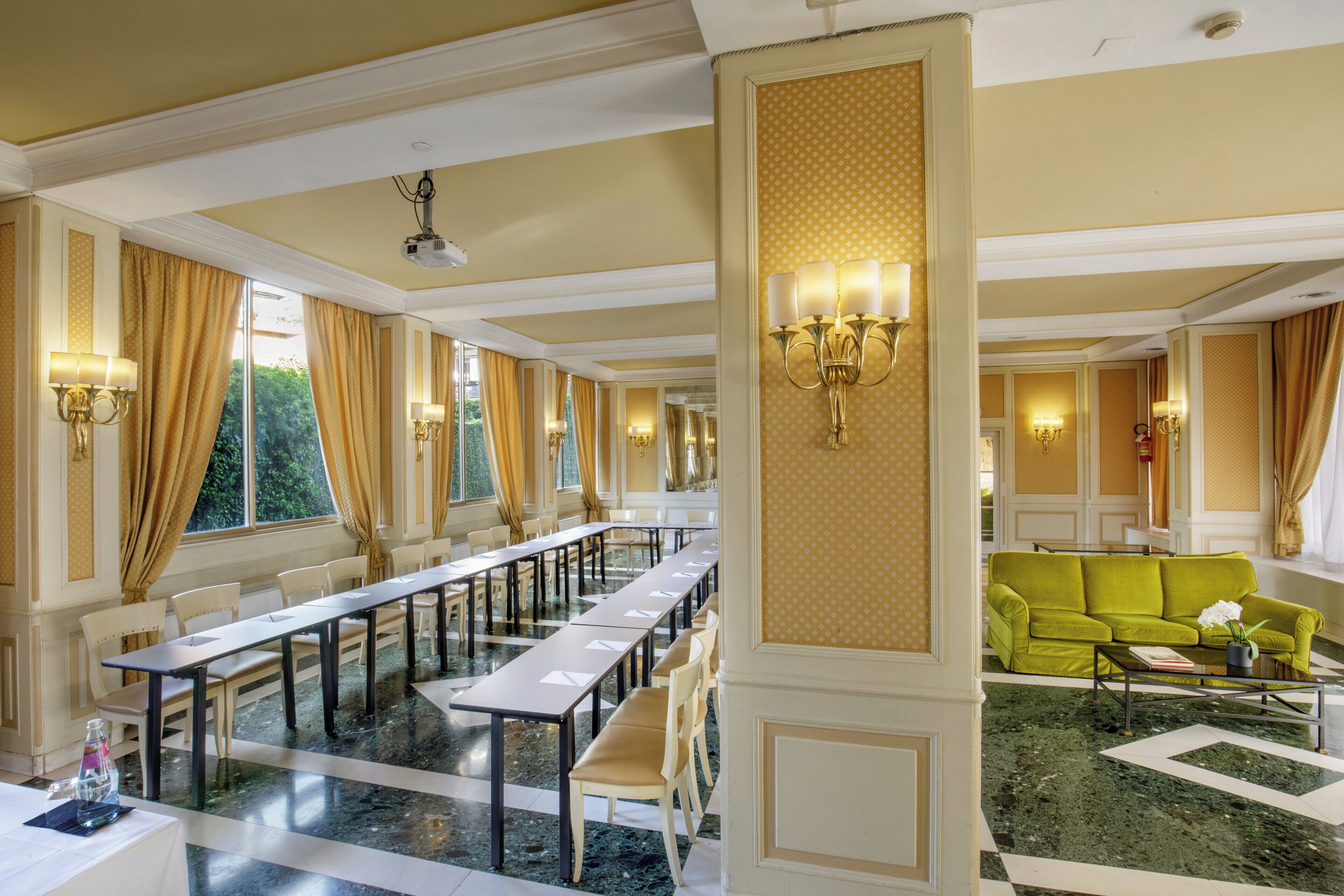Un hall d’hôtel avec une longue table à manger, de nombreuses chaises, un canapé vert et un mur orné de motifs.