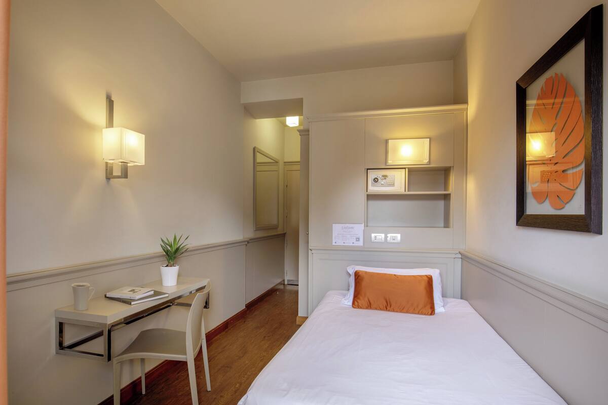 A hotel room with a bed, a desk, a chair, a potted plant, a framed artwork, and a wall-mounted shelf.