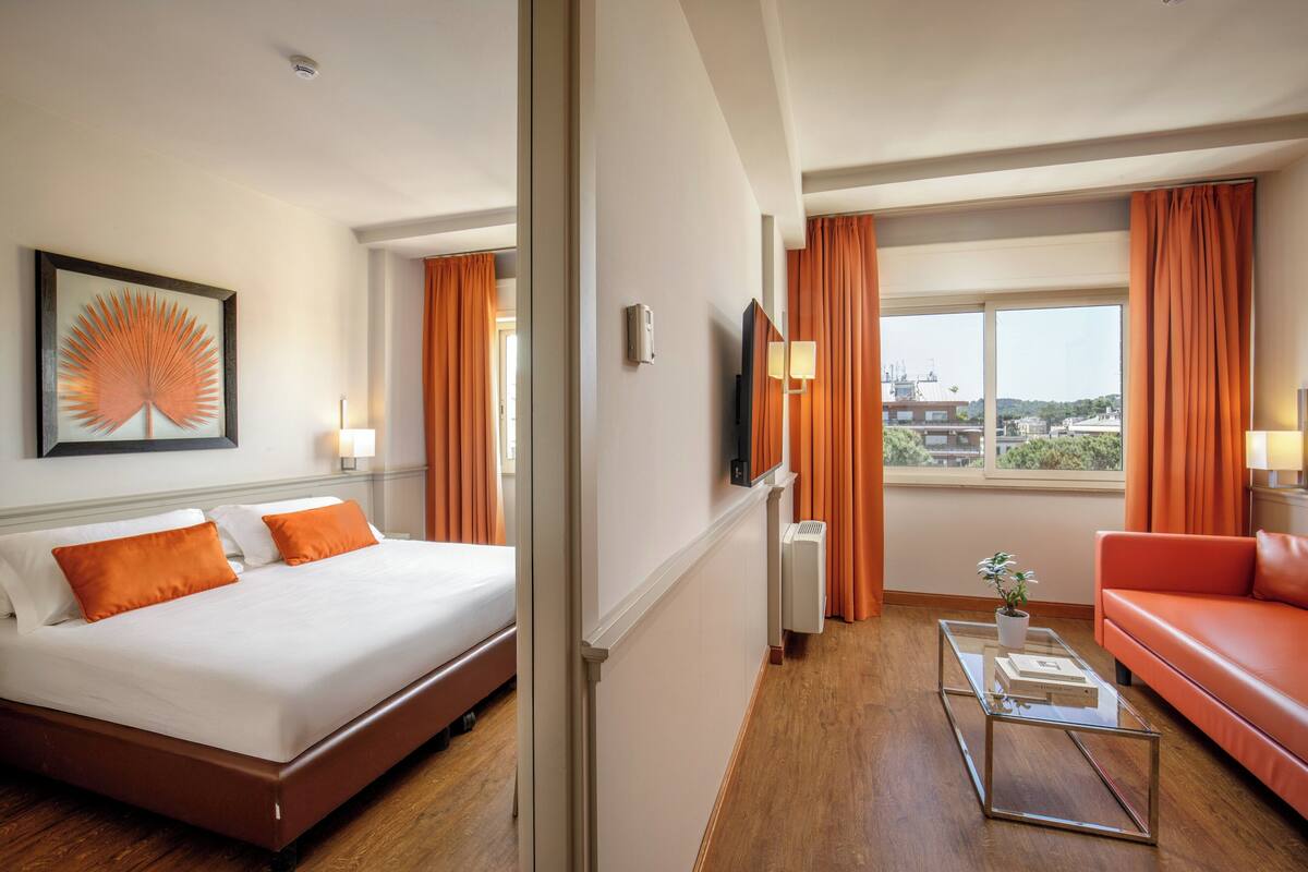 A hotel room with a bed, a sofa, a coffee table, a TV, and a window with curtains.