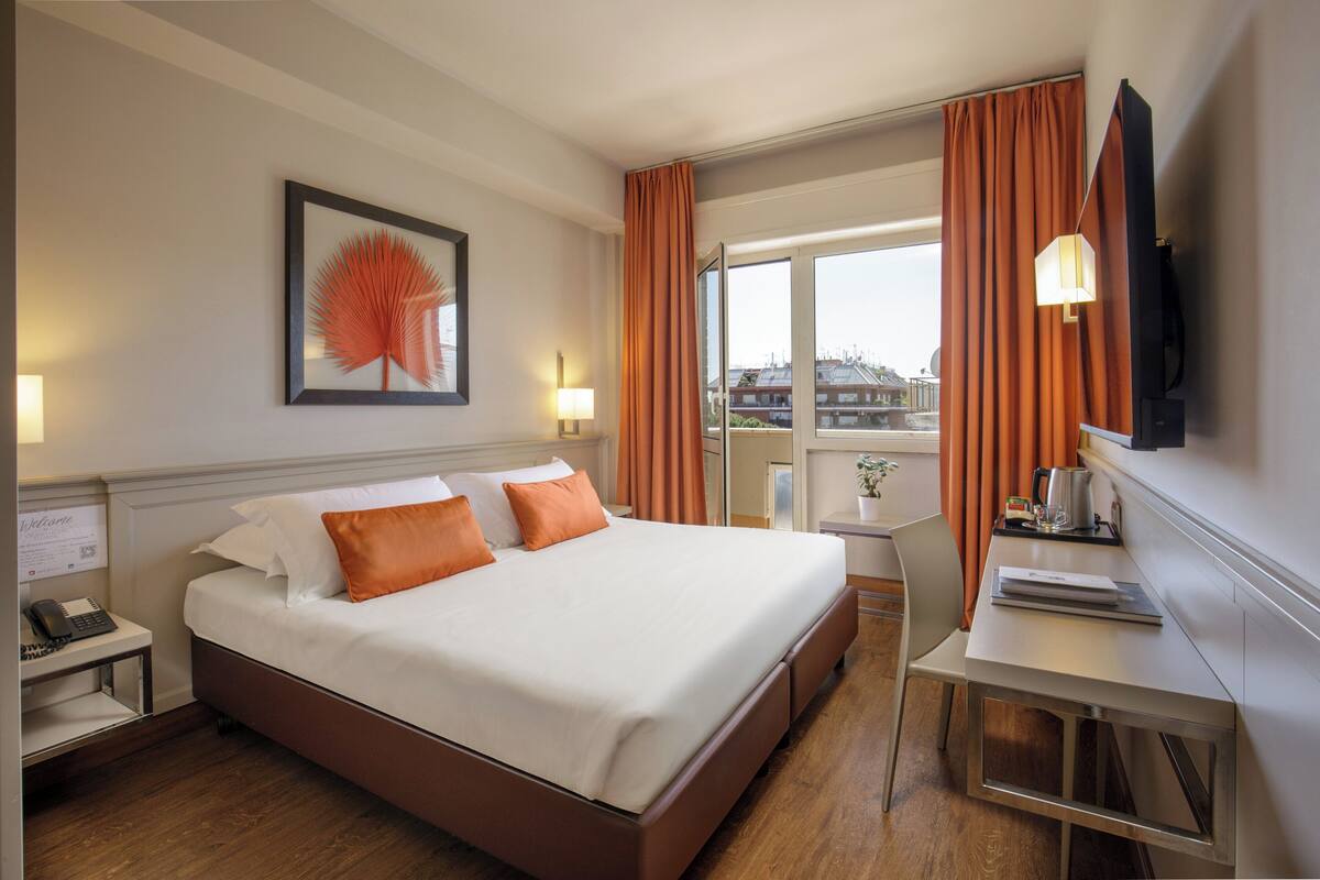 A hotel room with a large bed, a desk, a chair, a TV, and a window with curtains.
