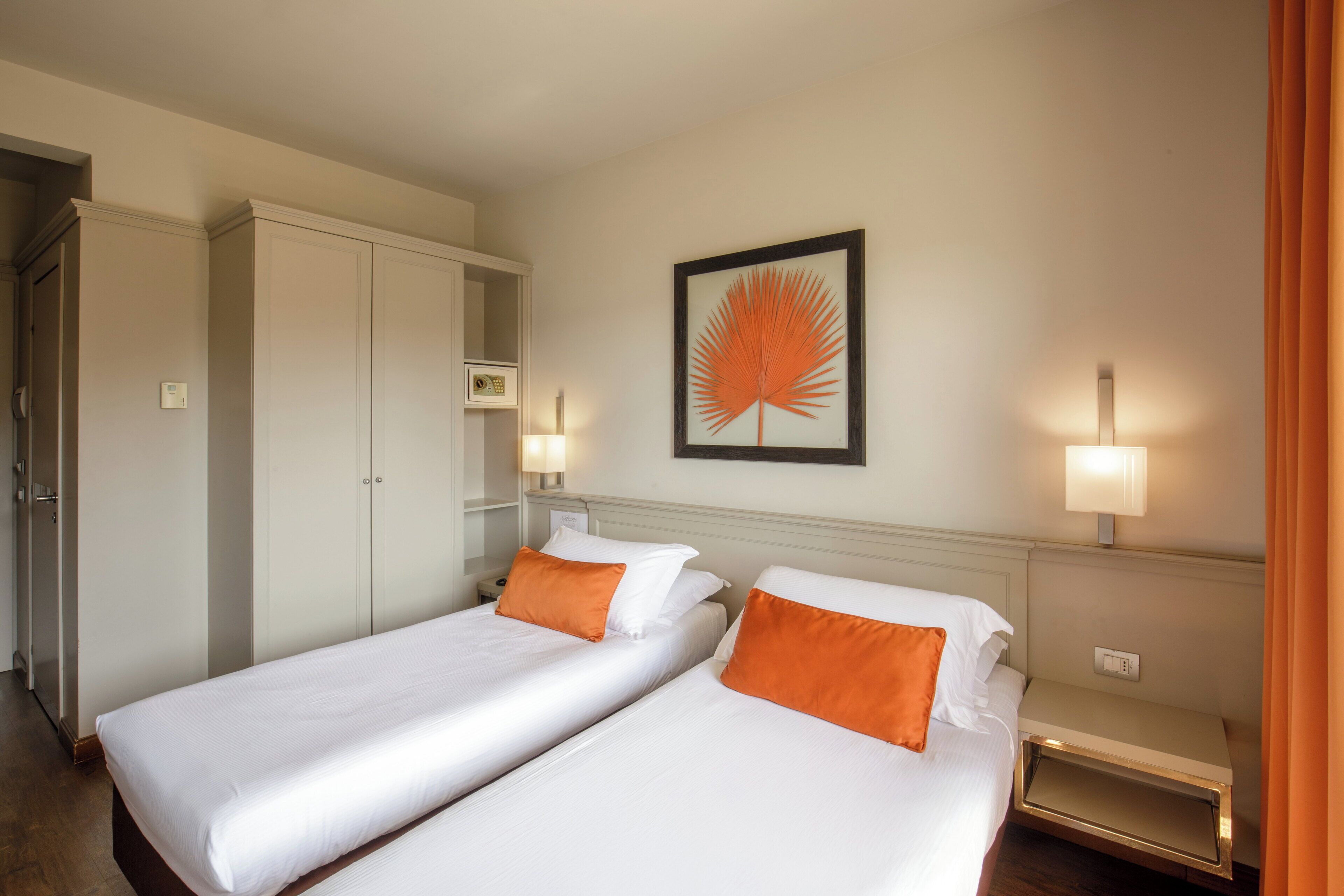 Habitación de hotel con dos camas individuales, un armario, una mesita de noche, una obra de arte enmarcada y una lámpara de pared.