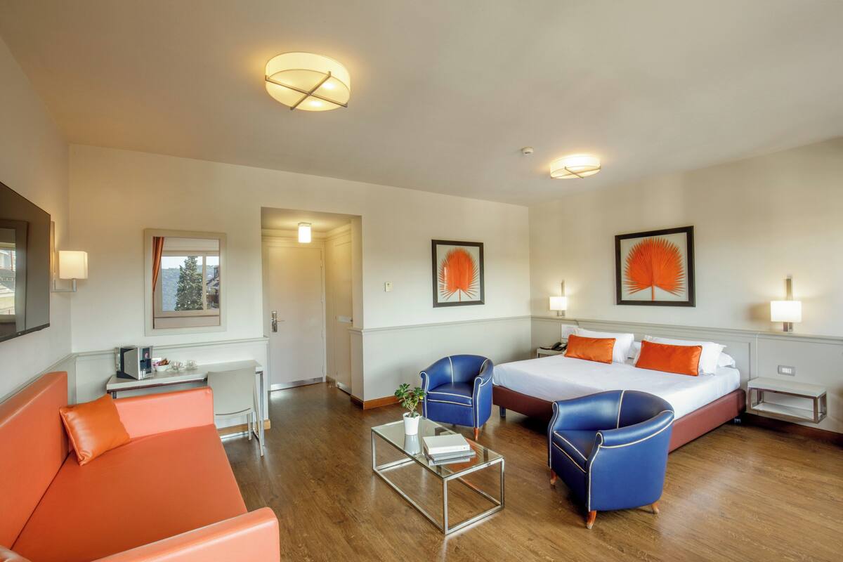 A hotel room with a bed, two blue armchairs, a small table, and a desk with a lamp.