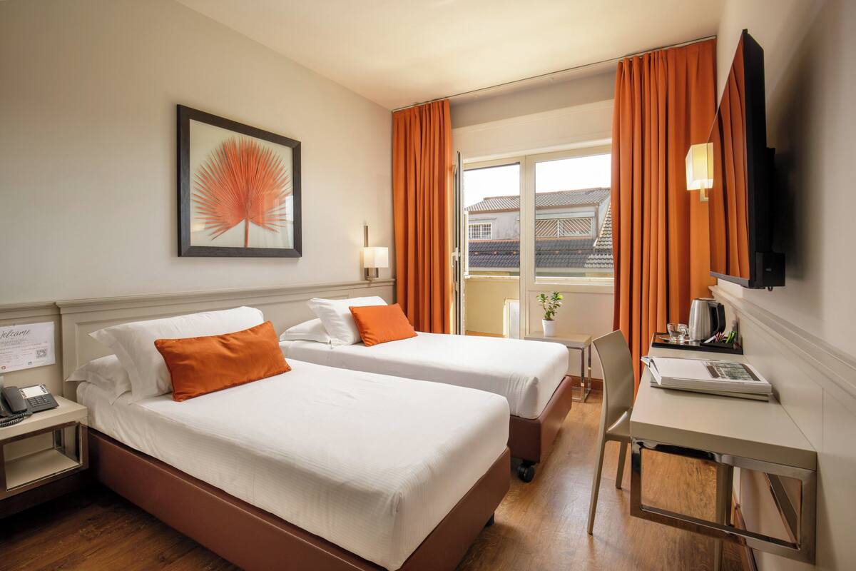 A hotel room with two beds, a desk, a chair, a TV, and a window with curtains.