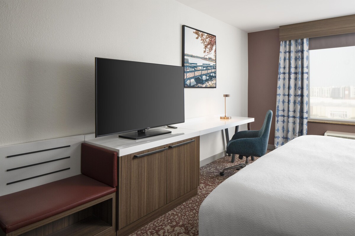 A hotel room with a bed, a TV on a desk, a chair, a desk with drawers, a window with curtains, and a painting on the wall.