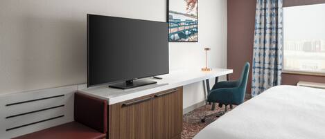 Habitación de hotel con cama, televisor sobre un mueble de madera, escritorio con silla y ventana con cortinas.
