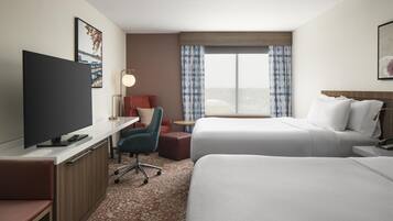 A hotel room with a flat-screen TV, a desk with a lamp, two beds, a chair, and a window with curtains.