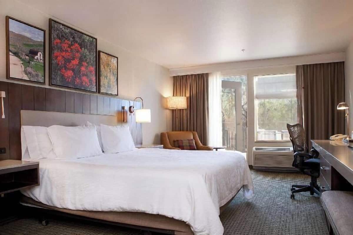 A hotel room with a large bed, a desk with a chair, a lamp, and a window with curtains.