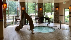 A spa area with a hot tub, stone pillars, wicker chairs, and large windows with views of greenery.