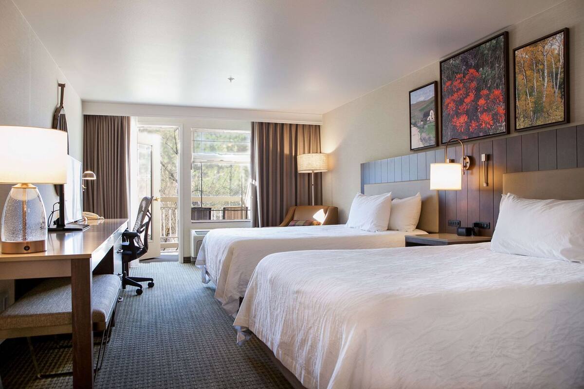 A hotel room with two beds, a desk, a chair, a lamp, a phone, and a window with curtains.