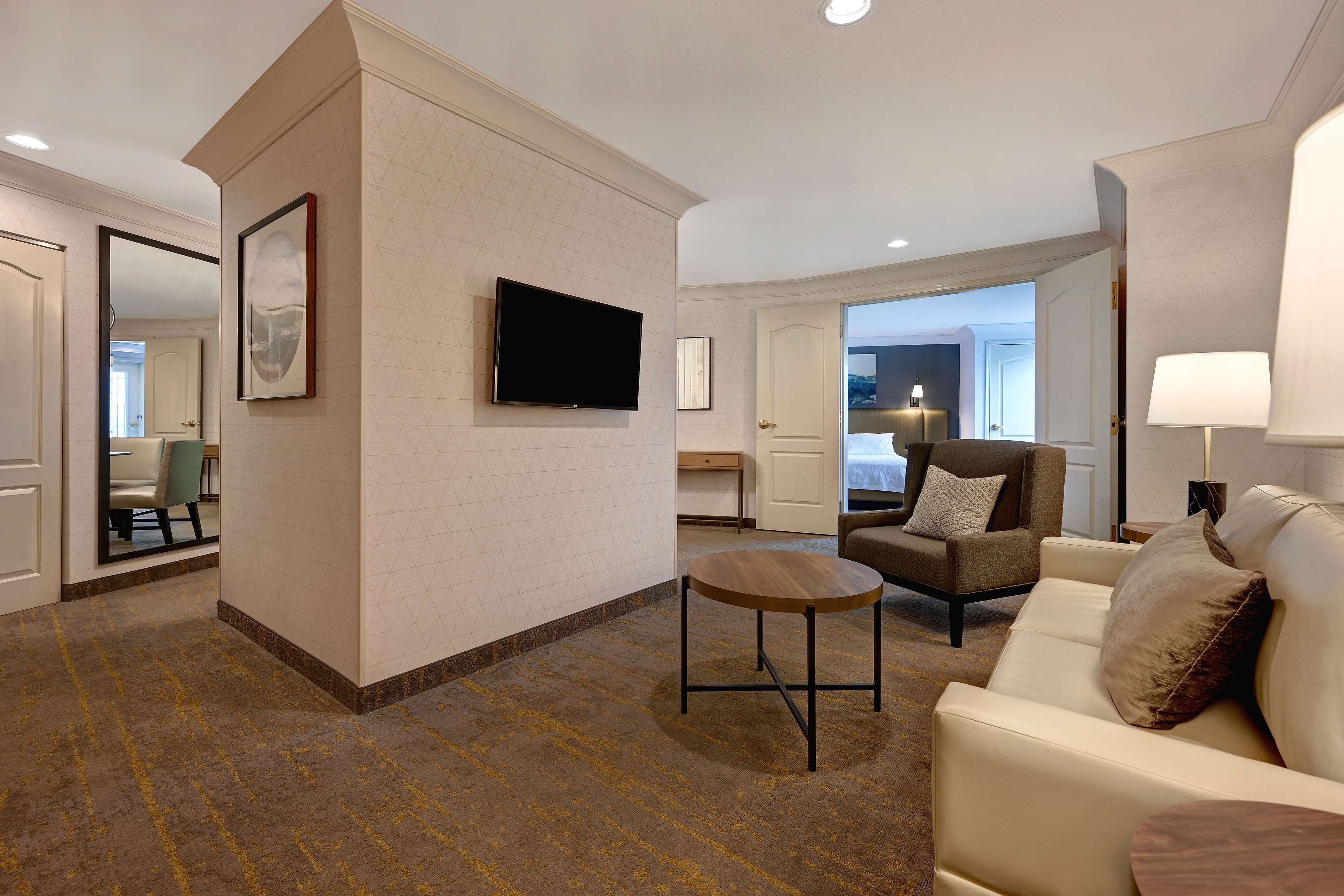 A hotel room with a sofa, armchair, and a small table. There is a television mounted on the wall and a door leading to another room.