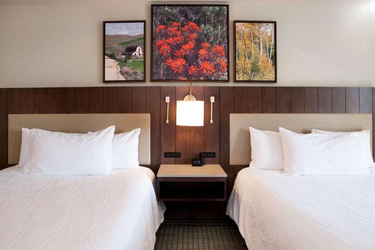 A hotel room with two beds, a nightstand, a lamp, and three framed pictures on the wall.