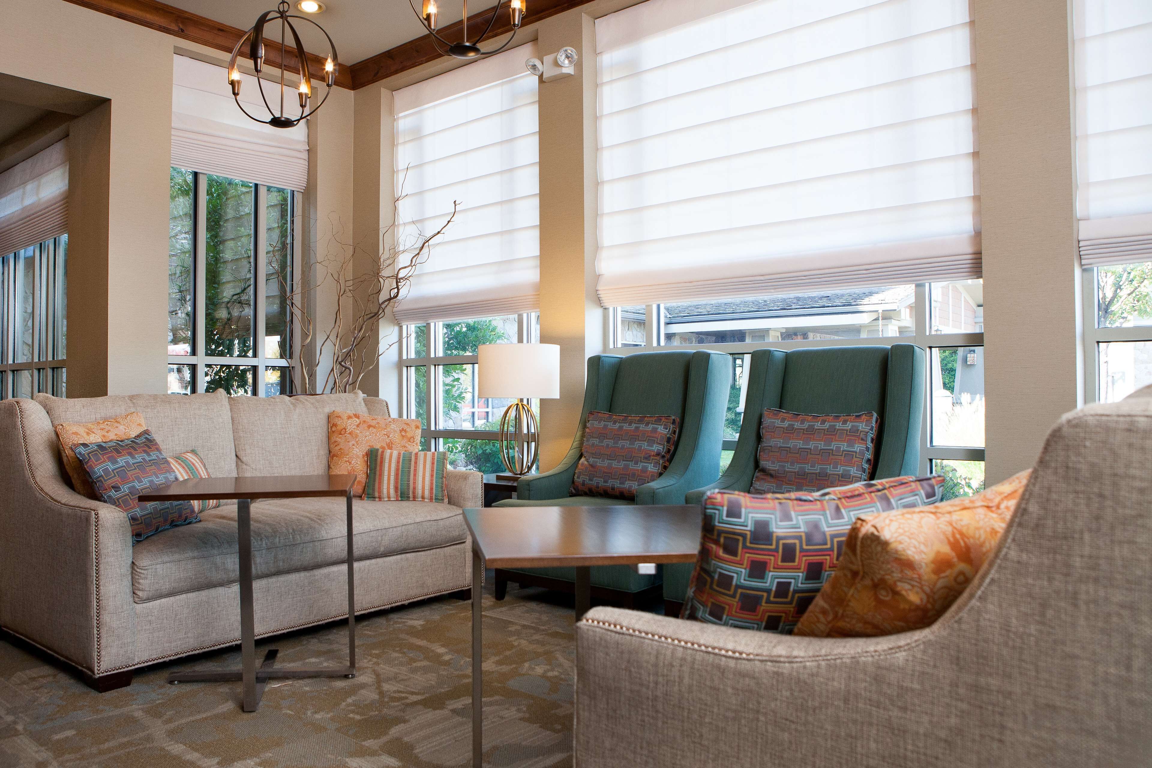 A spacious living room with a sofa, armchair, and a small table. Large windows with blinds allow natural light to fill the room.