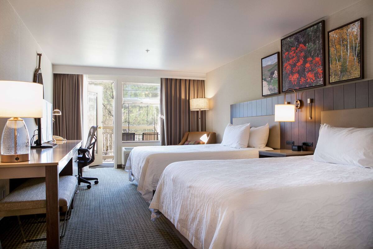 A hotel room with two beds, a desk with a lamp, a chair, and a view of the outdoors.