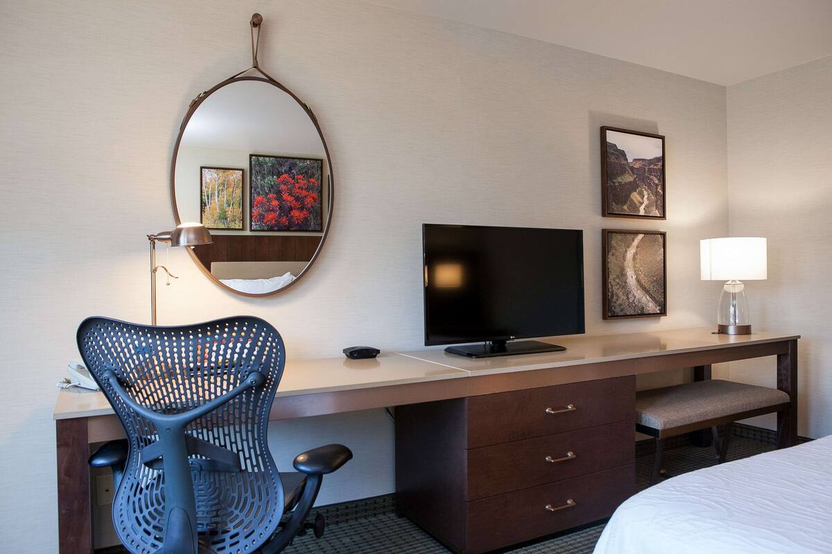 A hotel room with a desk, a chair, a television, a bed, a lamp, and two framed pictures on the wall.