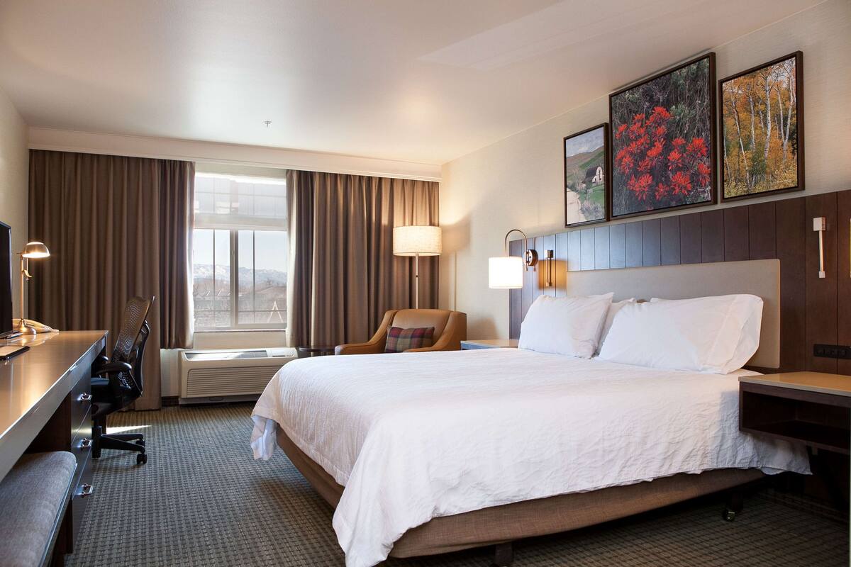 A hotel room with a large bed, a desk with a chair, a lamp, and a window with curtains.