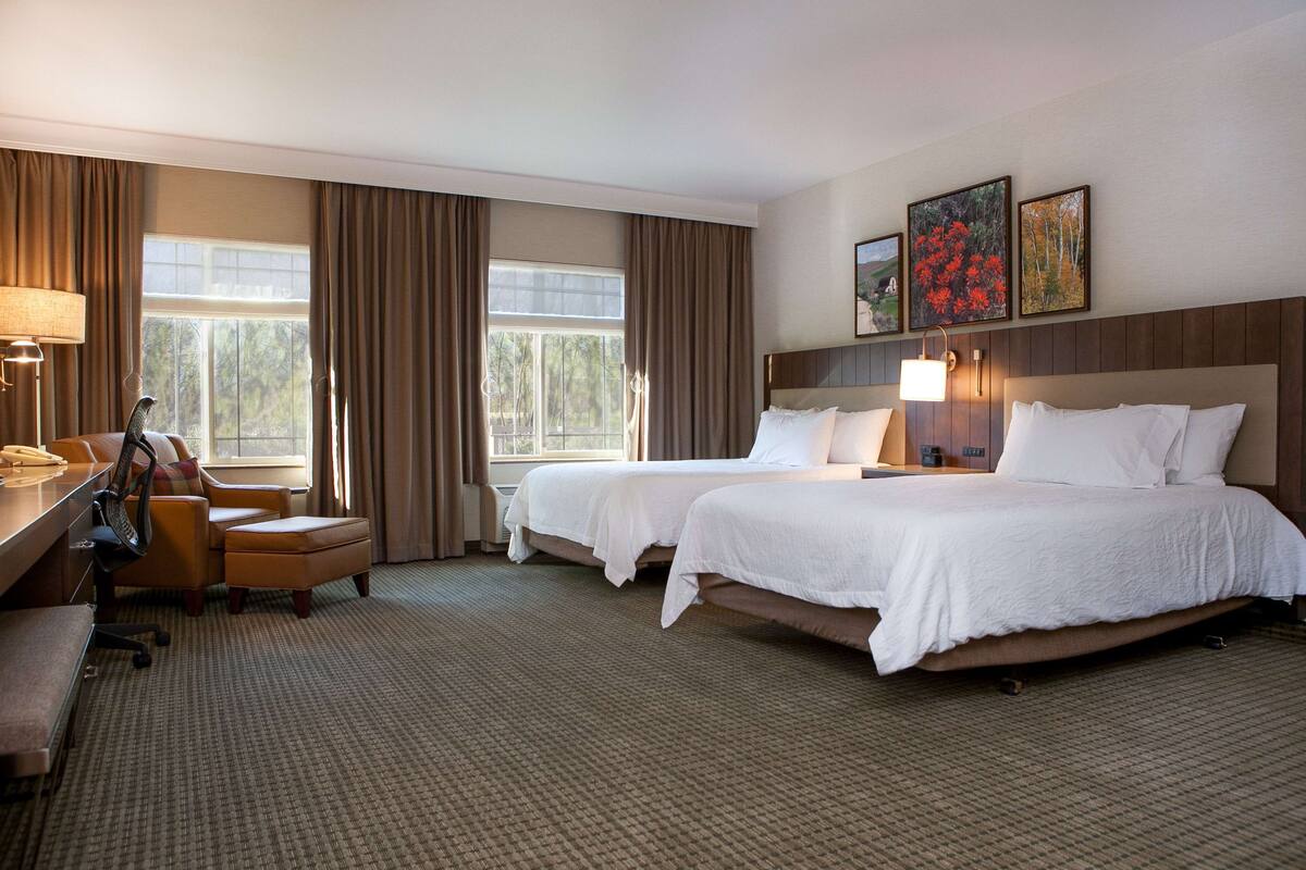 A hotel room with two beds, a desk, a chair, and a window with curtains.