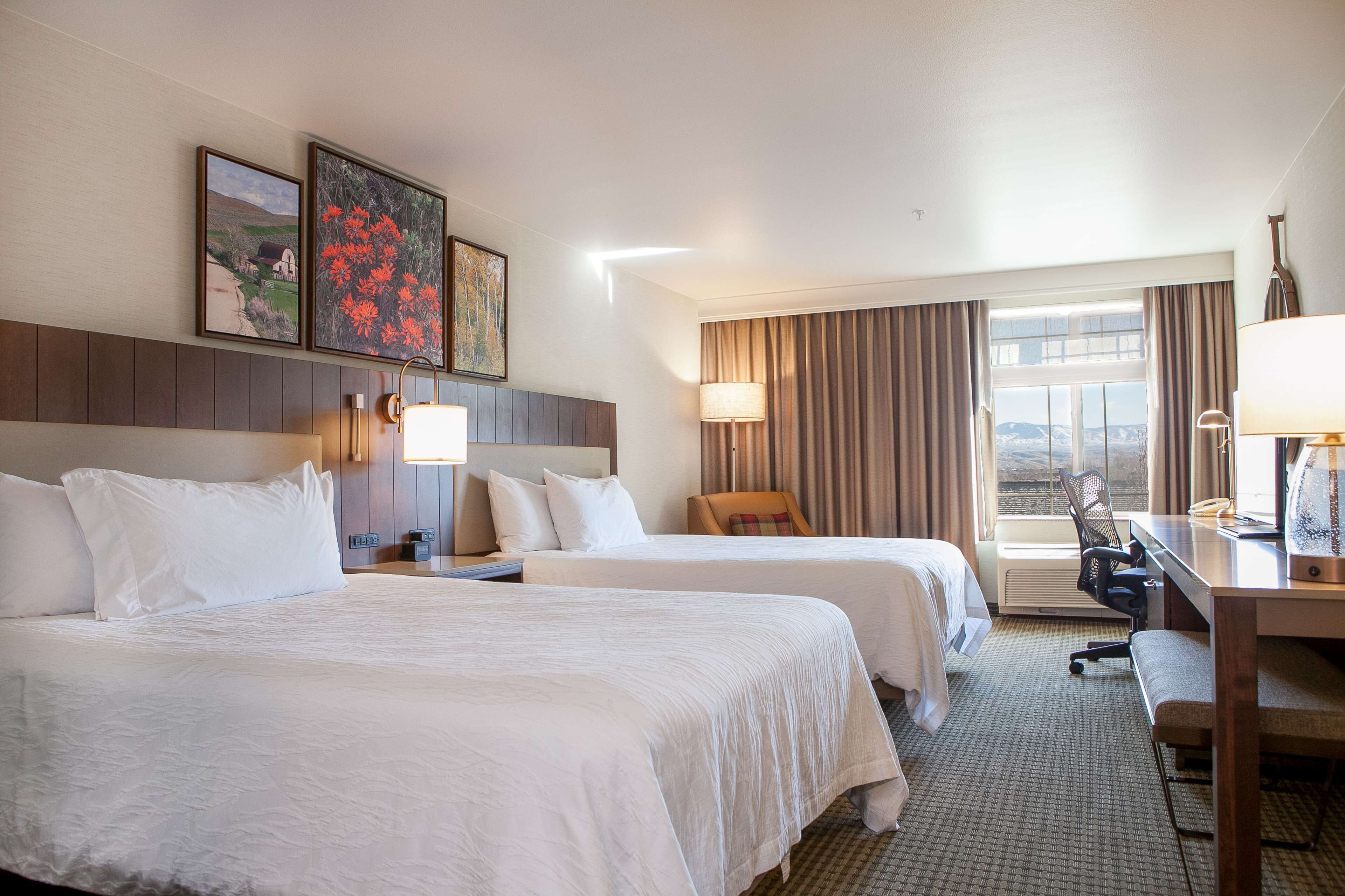 A hotel room with two beds, a desk, a chair, and a large window with curtains.