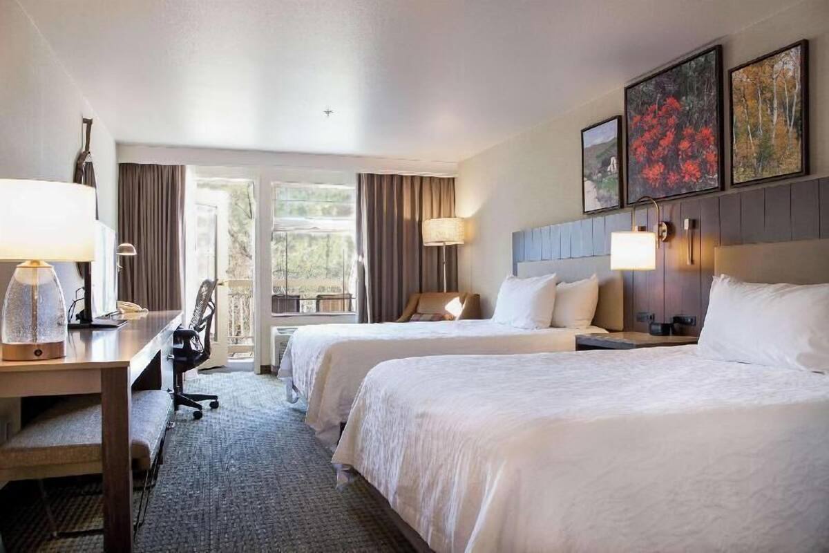A hotel room with two beds, a desk with a lamp, a chair, and a large window with curtains.