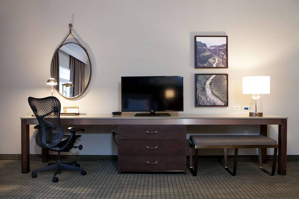 A hotel room with a desk, chair, TV, lamp, and two framed pictures on the wall.