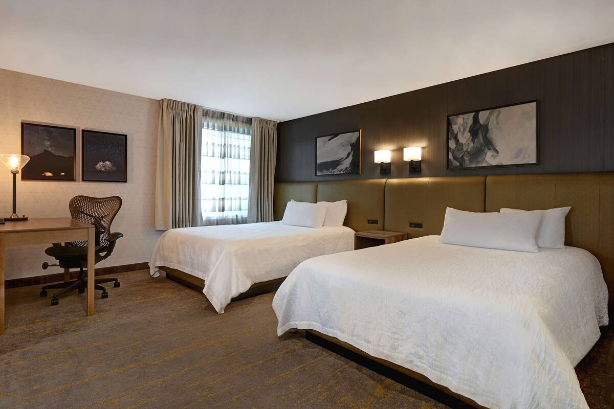 A hotel room with two beds, a desk, a chair, and artwork on the walls.