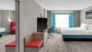 A hotel room with a bed, a desk with a TV, a chair, a bench, and a window with curtains.
